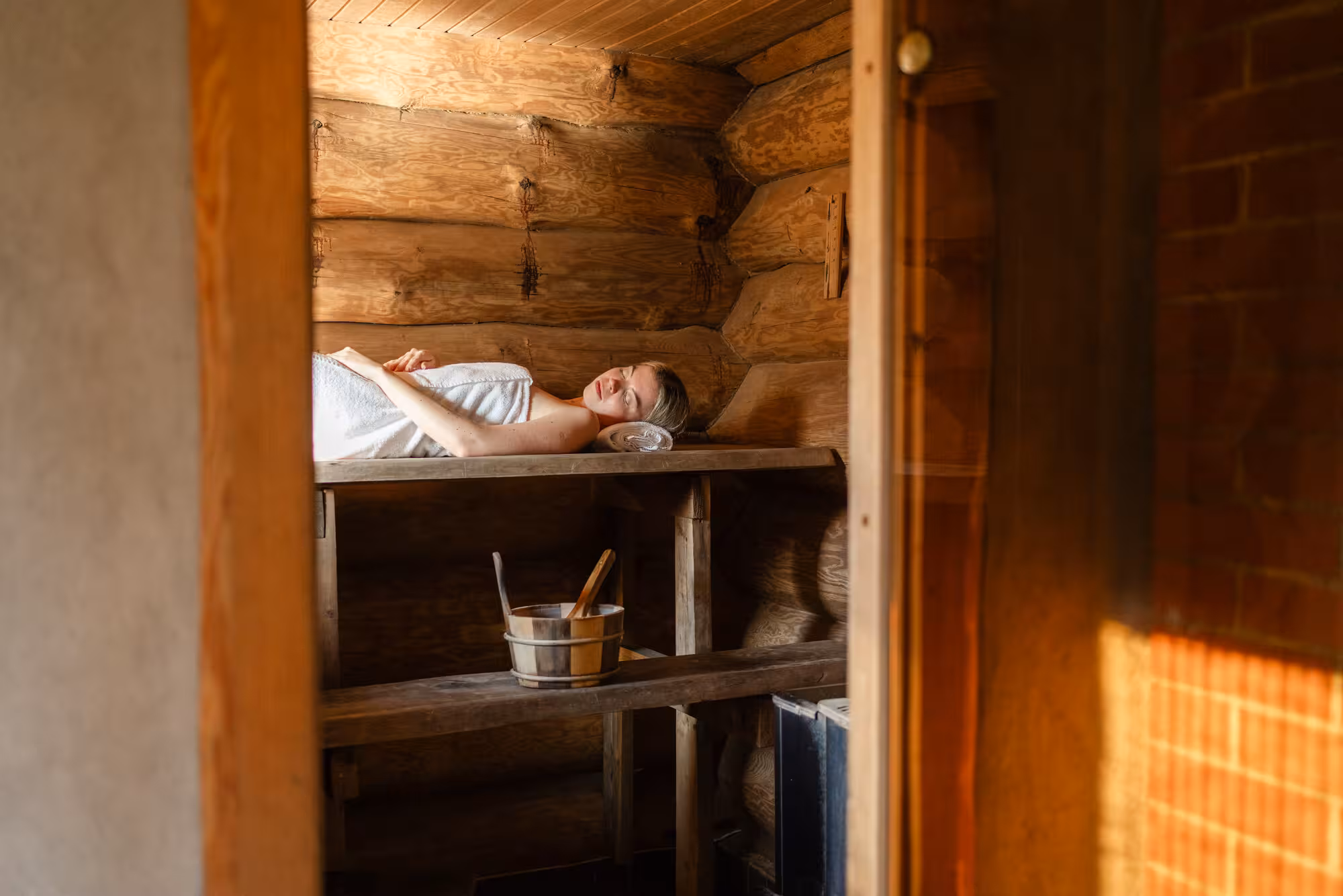 Valges rätikus naine pikutamas traditsioonilise puidust palksauna laval, kus alumisel astmel on näha puidust saunakibu.