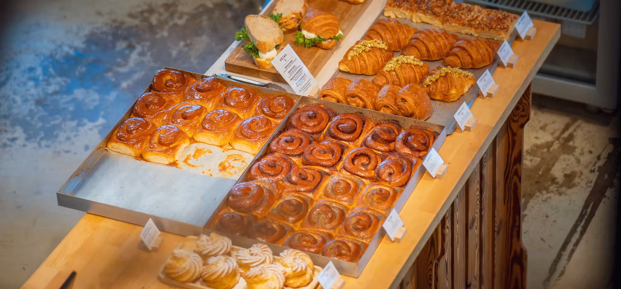 Mitmekülgne valik värskeid küpsetisi, sealhulgas kaneelisaiakesi, croissante ja võileibu, eksponeerituna pagariäri puidust letil.